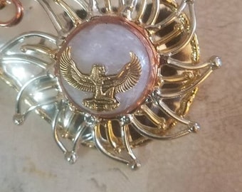 May include: Gold and silver toned pendant featuring a white stone with a gold winged figure in the centre. The pendant is surrounded by a decorative, sunburst-like design.