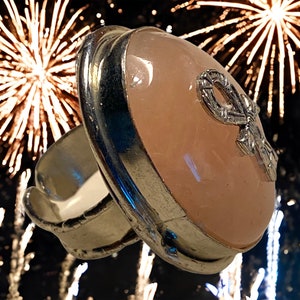 May include: A silver ring with a large oval-shaped pink gemstone. The gemstone has a silver design on it. The ring is photographed against a background of fireworks.