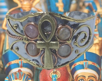 May include: A silver ring with a gold ankh symbol and five pink stones. The ring is decorated with a swirling design.