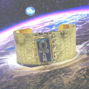 May include: Gold cuff bracelet featuring an ankh symbol set in blue and green enamel. The bracelet is decorated with hearts, arrows, and the word "LOVE".