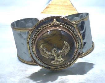 May include: A wide silver cuff bracelet with gold and bronze accents. The centerpiece features a round, tiger's eye stone with a gold hieroglyphic design. The bracelet has a hammered texture and an ancient Egyptian theme.