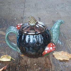 Puede incluir: Una tetera de cerámica hecha a mano con un acabado oscuro y brillante. La tetera presenta una tapa y un asa en forma de hongo, con detalles de hongos rojos y blancos. El pico y el asa son de color verde apagado.