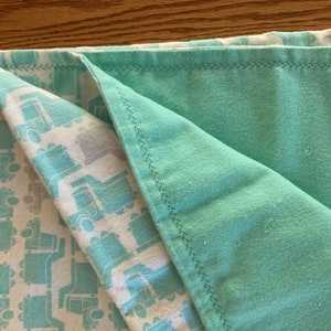 May include: A folded baby blanket with a teal and white pattern of trucks on one side and a solid teal color on the other. The edges are sewn with a black thread.