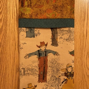 May include: A rectangular fabric item with a fall theme. The top section features a brown fabric with autumn leaves. Below is a green band, and the main section displays a rural scene with scarecrows and a farmhouse.