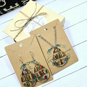 May include: A set of book-themed jewelry featuring a necklace and earrings. The pendant and earrings are teardrop-shaped and filled with colorful books on a bookshelf. The necklace is silver and has a simple chain. The earrings are silver with a hook closure. The jewelry is packaged on brown cardstock with the text "Sparrow & The Tangled Sparrow Handmade Jewelry".
