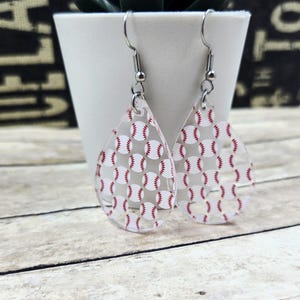 May include: Clear acrylic teardrop earrings featuring a pattern of small baseballs with red stitching. The earrings have silver-colored fish hook ear wires. The background includes a white pot and a wooden surface.