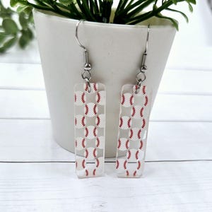 May include: A pair of silver earrings with clear rectangular charms featuring a repeating pattern of white baseballs with red stitching.