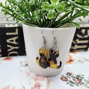 May include: A pair of cat-shaped earrings with a tortoiseshell pattern. The earrings are made of a translucent material with shades of brown and yellow. They hang from silver-colored hooks.