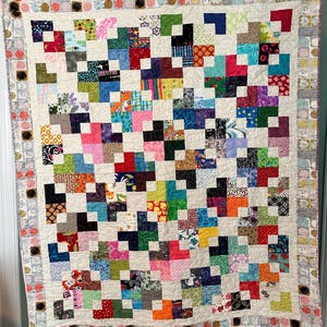May include: A patchwork quilt featuring a grid of multi-colored fabric squares in various patterns, including floral, geometric, and abstract prints. The quilt is bordered by a strip of light-colored fabric with circular motifs.