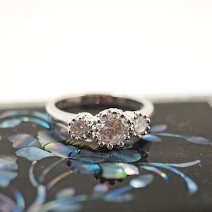 May include: A silver ring with three clear gemstones, set on a black box with floral mother-of-pearl inlay. The ring features three round, sparkling gemstones. The box has a decorative border, and the inlay has a floral design.