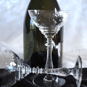 Könnte beinhalten: Zwei klare, antike Champagner-Coupé-Gläser mit geätzten Blumendesigns, eines aufrecht und eines auf der Seite. Im Hintergrund befindet sich eine grüne Champagnerflasche, die ein festliches Thema suggeriert.