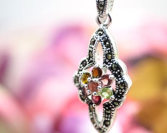 May include: A silver pendant with a floral design, featuring a cluster of colorful gemstones. The pendant hangs from a delicate silver chain. The gemstones include red, green, and brown stones.