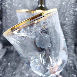 May include: Clear wine glass with a gold rim and etched floral and shell designs. The glass has a stem and is partially filled. A wine bottle is visible in the background. The glass is elegant and decorative.