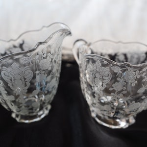 Könnte beinhalten: Ein Set aus Sahne- und Zuckerdose aus klarem Glas. Beide Teile weisen ein Blumen- und Scrollmuster auf, das in das Glas geätzt ist. Der Sahneaufgießer hat einen Ausguss und einen Griff, während die Zuckerdose einen Griff auf jeder Seite hat.