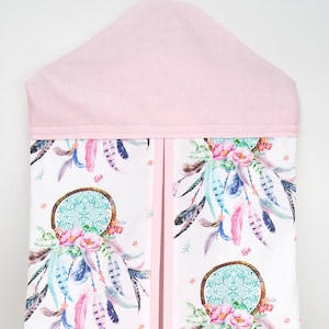 May include: A pink fabric diaper caddy with a white background featuring a repeating pattern of dreamcatchers, feathers, and flowers. The caddy has a pink fabric top and a hanger.