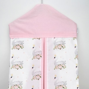 May include: A pink and white diaper caddy with a swan and floral print. The caddy has a pink flap on top and hangs from a hook.