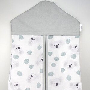 May include: A hanging nursery organizer with a gray top and a white body. The body is patterned with koala bear faces and eucalyptus leaves in shades of gray and blue-green. The organizer has two vertical sections with gray trim.