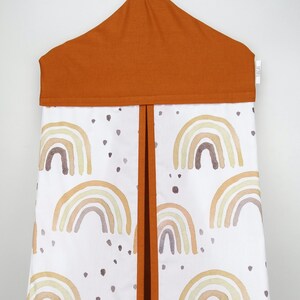 May include: A white diaper caddy with a brown top and a pattern of colorful rainbows and brown dots. The caddy has a flap that can be opened and closed to access the contents.