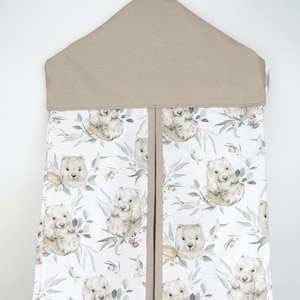 Puede incluir: Un organizador de pañales de tela beige con un fondo blanco que presenta un patrón repetitivo de ilustraciones de acuarela de wombats en un bosque de eucaliptos. El organizador tiene un lazo para colgar y un divisor en el centro.