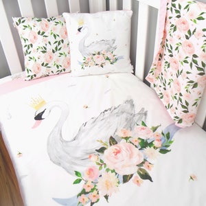 May include: A white crib with a pink and gray floral patterned bedding set. The bedding features a white swan with a gold crown and a floral design. The crib also has a pink and gray floral patterned pillow and a pink and gray floral patterned blanket.