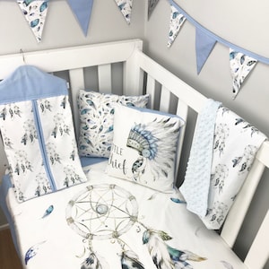 May include: A white cot with blue and white bedding featuring a dreamcatcher design and feathers. The bedding also includes a pillow with the text "Little Chief" and a headdress design. A blue and white bunting hangs above the cot.