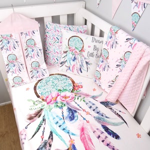 May include: A pink and white crib bedding set with a dreamcatcher design. The set includes a fitted sheet, a crib skirt, a diaper stacker, a pillow, and a blanket. The dreamcatcher is decorated with feathers, flowers, and beads. The text "Dream Big Little One" is printed on the pillow.