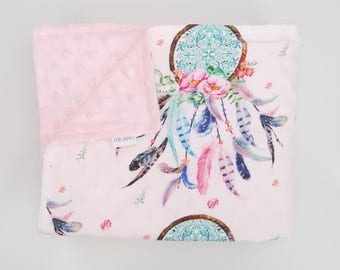 Puede incluir: Manta de bebé rosa y blanca con un diseño de atrapasueños con plumas, flores y un mandala turquesa. La manta tiene una textura suave y afelpada.