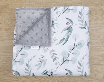 Puede incluir: Una manta blanca para bebé con un tejido minky gris con puntos en un lado y un estampado de acuarela de hojas y ramas de eucalipto en el otro lado. La manta tiene la palabra "DANNOH" impresa en ella.