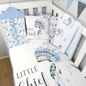 Puede incluir: Un juego de ropa de cama para cuna blanca con un patr&oacute;n de tocado azul y blanco. La ropa de cama incluye una s&aacute;bana bajera, una funda de almohada y una manta. El juego presenta el texto "Little Chief" en escritura negra.