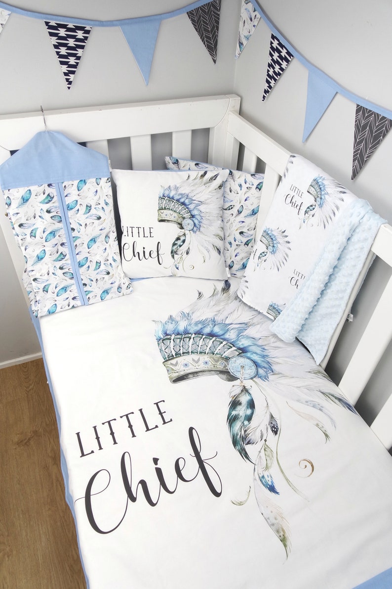 Puede incluir: Un juego de ropa de cama para cuna blanca con un patr&oacute;n de tocado azul y blanco. La ropa de cama incluye una s&aacute;bana bajera, una funda de almohada y una manta. El juego presenta el texto "Little Chief" en escritura negra.