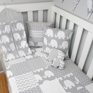 May include: A gray and white baby crib bedding set with a patchwork quilt featuring elephants, plus signs, and chevron patterns. The set includes a crib skirt, a pillow, and a stuffed elephant toy.