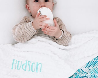 May include: A baby lying on a white and blue patterned blanket, drinking from a white bottle. The blanket has the name "Hudson" embroidered on it in turquoise.