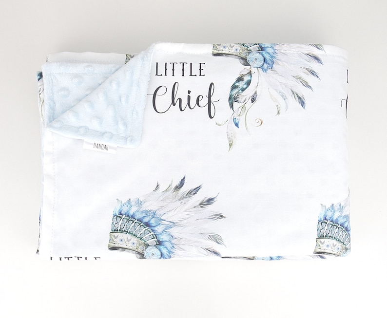 Puede incluir: Una manta de beb&eacute; blanca con un patr&oacute;n de tocado azul y blanco. La manta tiene las palabras "Little Chief" impresas.