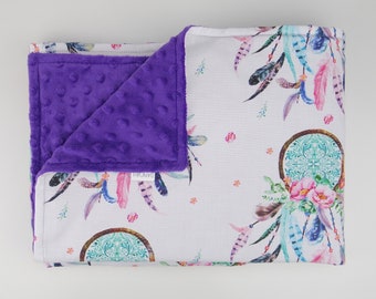 Puede incluir: Una manta de bebé blanca y morada con un patrón de atrapasueños y plumas. La manta es suave y acogedora, perfecta para una guardería o sala de juegos.