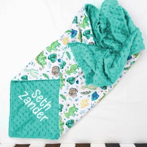 May include: A turquoise and white baby blanket with a dinosaur and tropical leaf print. The blanket is personalised with the name "Seth Zander" in white embroidery.