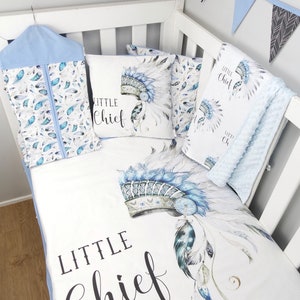 May include: A white cot bedding set with a blue and white headdress pattern. The bedding includes a fitted sheet, a pillowcase, and a blanket. The set features the text "Little Chief" in black script.