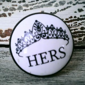 May include: White ceramic knob with a black crown and the word "HERS" in black lettering.