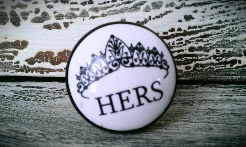 May include: White ceramic knob with a black crown and the word "HERS" in black lettering.