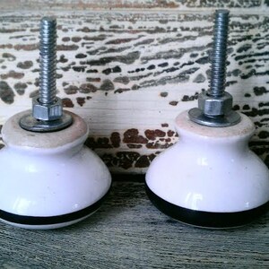 May include: Two white ceramic cabinet knobs with black trim and silver metal accents. The knobs have a screw-on design.