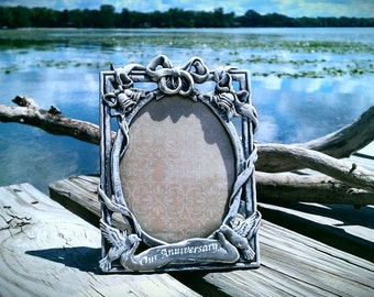 May include: A silver picture frame with an ornate design and a blank space for a photo. The frame has a white background with a floral pattern and the words "Our Anniversary" at the bottom.