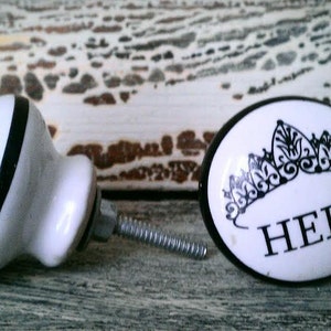 May include: Two white ceramic door knobs with black trim. One knob is round and features a crown design and the word "HERS". The other knob is round and features the word "HERS" in a black and white stripe design.