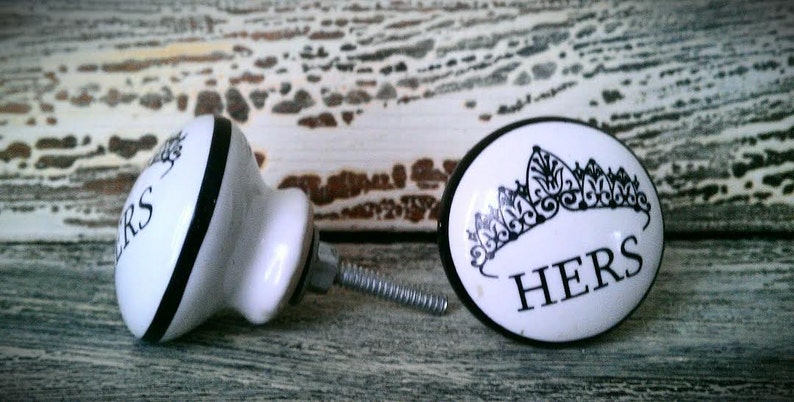 May include: Two white ceramic door knobs with black trim. One knob is round and features a crown design and the word "HERS". The other knob is round and features the word "HERS" in a black and white stripe design.