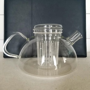 May include: A clear glass teapot with a built-in strainer. The teapot has a round body and a curved handle.
