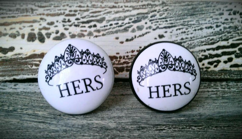 May include: Two white oval ceramic knobs with black crown and the word "HERS" printed on them.