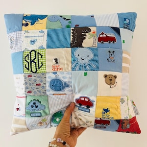 May include: A patchwork cushion with squares of fabric, each with embroidered images of dinosaurs, cars, an octopus, and a whale. The cushion has a light blue border. The cushion has the initials "SRC" embroidered on it.