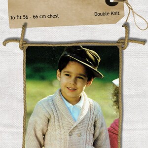 May include: A beige double-breasted knitted jacket for a child with a cable knit pattern. The jacket has a shawl collar and is fastened with buttons. The child is wearing a white shirt and a brown hat. The image includes the text "Childs Double Breasted Jacket" and "To fit 56 - 66 cm chest".