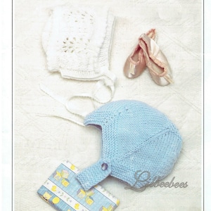 May include: Two knitted baby bonnets, one white with a lacy pattern and one light blue, with a small button on the top. The bonnets are laid out on a white surface with a small gift box wrapped in blue and yellow paper with a teddy bear and letter blocks design.