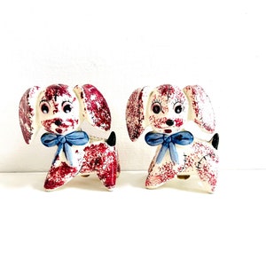 May include: A pair of ceramic dog salt and pepper shakers. The dogs are white with red and blue accents. They have blue bows around their necks and are standing on a white surface.