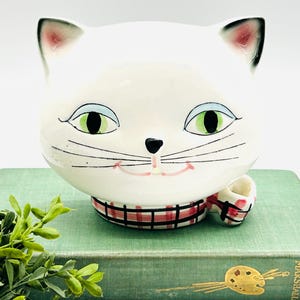 May include: A white ceramic cat head planter featuring large green eyes, blue eyelids, a small black nose, and painted whiskers. The cat wears a red and black plaid scarf around its neck, with a small matching plaid detail extending to the right side.