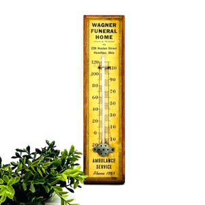 May include: A rectangular wall thermometer with a wooden frame and a yellow face. The text reads Wagner Funeral Home, 228 Heaton Street, Hamilton, Ohio, and Ambulance Service Phone 1953. It features a central glass tube and a metal bulb cover.